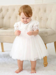 A baby girl wears a white knee-length lace christening dress with short sleeves and a tulle skirt.