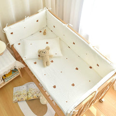 A white quilted crib bumper set with a small bear pattern and a matching pillow inside a wooden crib.