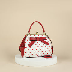 A white handbag with red polka dots, a red bow, and a gold kiss lock closure on a white pedestal.
