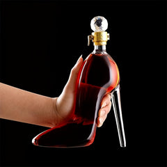 A hand holds a glass wine decanter shaped like a high-heeled shoe with a crystal-style stopper.