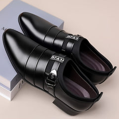 A pair of black pointed-toe men's formal shoes with a buckle strap and branded elastic detail.