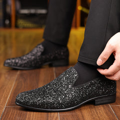 A pair of black pointed-toe slip-on dress shoes with a glittery finish worn with black trousers.