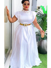 A woman wears a white sleeveless maxi dress with a high neck, pearl embellishments, and a gold belt.