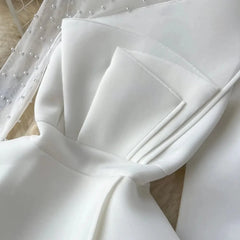 A close-up of a white mini dress featuring a pleated bodice and sheer pearl-embellished sleeves.