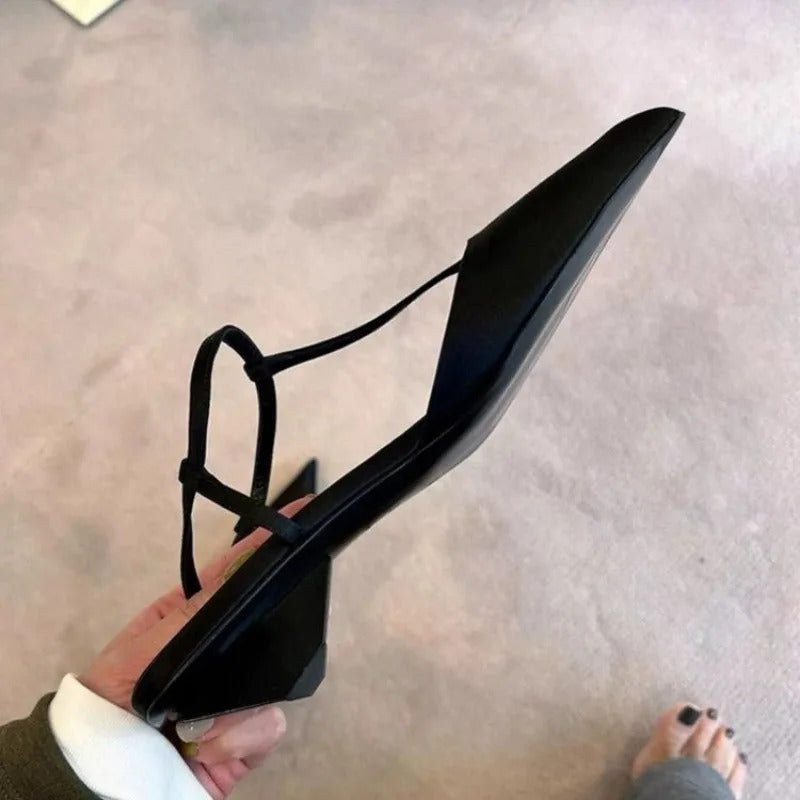 A hand holds a black pointed-toe sandal with a low chunky heel and thin ankle strap.