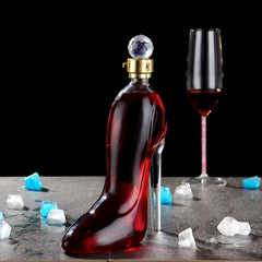 A high-heeled shoe shaped glass wine decanter filled with red wine sits on a table with ice cubes.