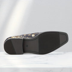 A black slip-on shoe with a gold floral pattern and a textured black sole sits on a marble surface.