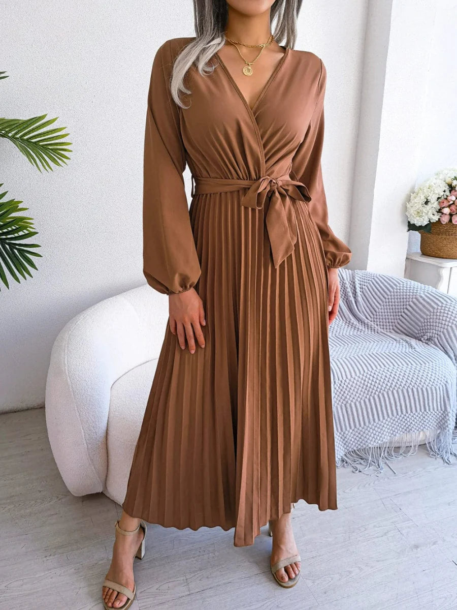 A woman wears a long-sleeved, brown pleated maxi dress with a V-neck and a tie-waist belt.
