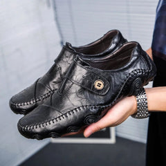 A pair of black leather slip-on loafers with a decorative button strap and textured rubber soles.