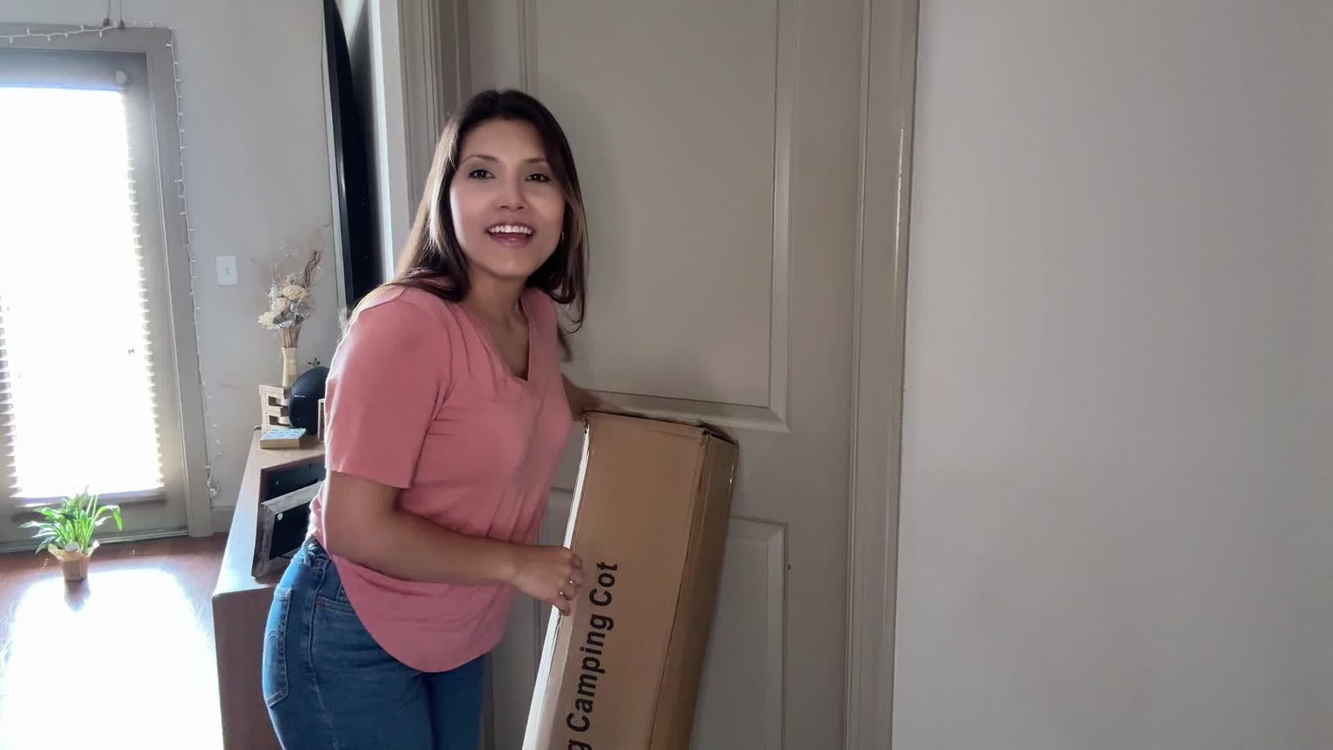 A woman stands next to a tall, rectangular cardboard box labeled as a folding camping cot.