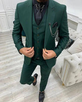 A man wears a dark green three-piece slim fit suit with a black shirt and silver chain accessory.
