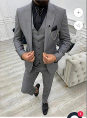 A man wears a slim-fit grey three-piece suit with a black shirt, tie, and decorative lapel chain.