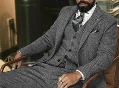 A man wears a gray herringbone wool three-piece suit with a white shirt and a patterned tie.