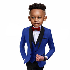 A young boy wears a formal three-piece blue suit with a black shawl lapel and a red bow tie.
