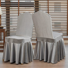 Two silver satin chair covers with a pleated skirt design displayed in a room with window blinds.