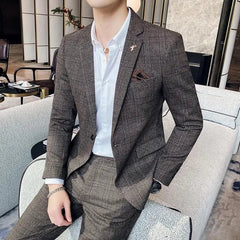 A man wears a brown plaid two-piece suit with a white button-down shirt and a lapel pin.