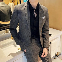 A man wears a grey plaid two-piece suit with a black button-down shirt and a white pocket square.