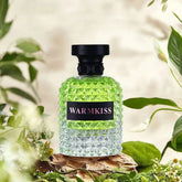 A bottle of Warmkiss perfume with a textured glass body and black cap sits on a wooden surface.