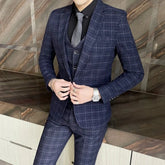 A man wears a navy blue plaid three-piece suit with a black shirt and grey tie.