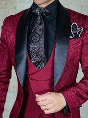 A man wears a burgundy patterned suit jacket with a black satin shawl lapel, vest, and tie.