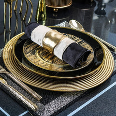 A black and gold glass dinner plate with a textured gold charger and a napkin in a gold ring.