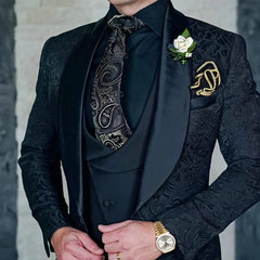 A man wears a black three-piece jacquard suit with a paisley tie and a gold pocket chain.