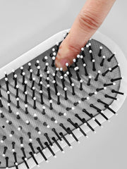 A finger presses into the cushioned base of a white airbag massage comb with black bristles.