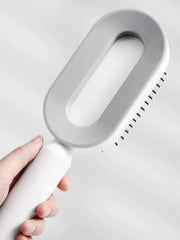 A hand holds a white and grey oval-shaped airbag massage comb with a long handle.