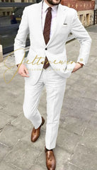 A man wears a white two-piece suit with a patterned tie and brown leather loafers.