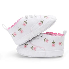 A pair of white canvas baby sneakers featuring pink flower embroidery and a soft sole.