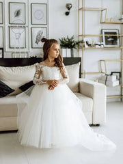 A young girl wears a white floor-length ball gown with long lace sleeves and a boat neckline.