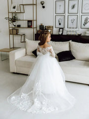 A young girl wears a white floor-length ball gown with long lace sleeves and a tulle train.