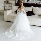 A young girl wears a white floor-length ball gown with long lace sleeves and a floral train.