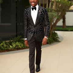 A man wears a black floral patterned tuxedo jacket with velvet accents and matching trousers.