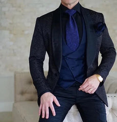 A man wearing a black patterned tuxedo jacket with a navy blue vest, tie, and black trousers.