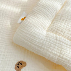 A cream-colored muslin baby blanket featuring a small embroidered bear and a soft quilted texture.