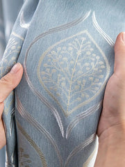 A pair of hands holds a blue curtain featuring a textured, embroidered leaf pattern.