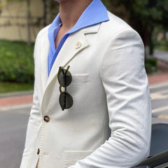 A man wears a white seersucker blazer with a blue collared shirt and sunglasses in the pocket.