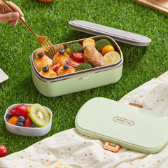A light green thermal lunch box with a stainless steel interior sits on grass with food inside.