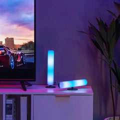 Two cylindrical RGB light bars with blue and white glow sit on a white cabinet next to a TV.