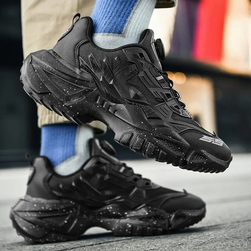 A person wears lace-free men's luxury sneakers with textured soles and white speckle details, paired with blue socks and beige pants, standing outdoors on a city street.