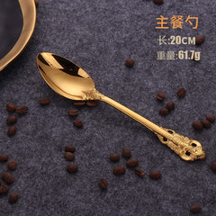 A gold-colored stainless steel dinner spoon with ornate relief detailing on the handle, displayed on a dark textured surface with scattered coffee beans.