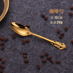 A gold-colored stainless steel coffee spoon with an ornate relief pattern on the handle, displayed on a dark textured surface with scattered coffee beans.