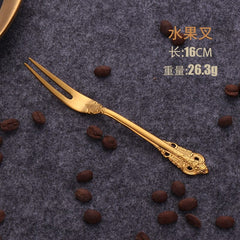 A gold-colored stainless steel fruit fork with an ornate relief handle design.