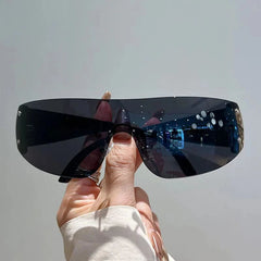 A hand holds a pair of rimless Y2K-style sunglasses with a single dark, mirrored wraparound lens.