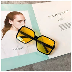Oversized square sunglasses with black frames and yellow lenses resting on a magazine.