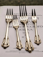Four ornate gold-toned stainless steel forks with intricate relief patterns on the handles.