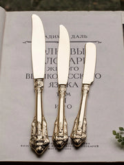 Three gold-toned stainless steel knives with ornate relief handles rest on a vintage book.