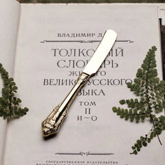 A gold-toned stainless steel butter knife with an ornate, vintage-style relief handle.