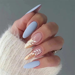 A hand displays almond-shaped press-on nails featuring matte blue polish and gold leaf leaf art.